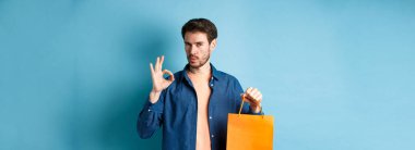 Yakışıklı genç adam dükkan tavsiye ediyor, turuncu alışveriş çantası ve tamam işareti gösteriyor, mavi arka planda duruyor..