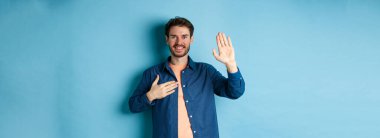 Mutlu genç adam kendini tanıtıyor. Elleri yüreğinde ve dostça gülümseyerek mavi arka planda durarak gerçeği söylüyor..