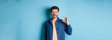 Olumlu gülümseyen adam göz kırpıyor ve iyiye işaret gösteriyor, iyi işi övüyor, iyi bir arkadaş tavsiye ediyor, mavi arka planda duruyor..