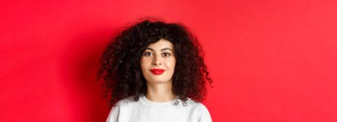 Kıvırcık saçlı, kırmızı dudaklı, kameraya mutlu gülümseyen, stüdyo arka planında duran güzel, modern bir kadının portresini yakından çek..