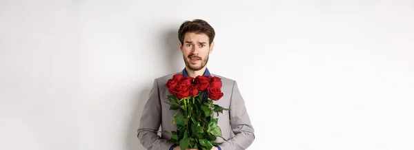 stock image Worried boyfriend in suit, holding flowers roses and looking doubtful at camera, standing with bouquet on valentines day against white background.