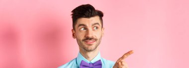 Bıyıklı ve papyonlu beyaz erkek modelin yakın çekimi, ilgi çekici bir gülümsemeyle solu işaret ediyor, tanıtım teklifini gösteriyor, pembe arka planda papyon takıyor..