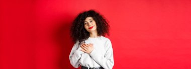 Kıvırcık saçlı, yumuşak bir genç kadın, kalbe sarılmış ve mutlu bir ifadeyle gülümsüyor, kırmızı arka planda dikilen, teşekkür edin..