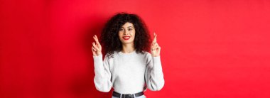 Kırmızı dudaklı, kıvırcık saçlı, şans dileyen ve dilek tutan umut dolu genç bir kadın. Hayallerinin gerçekleşmesi için dua eden heyecanlı, kırmızı arka planda gülümseyen..