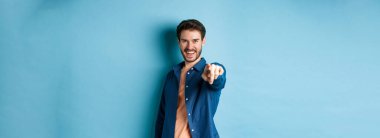 Kendinden emin gülümseyen adam seni seçiyor ya da davet ediyor, kameraya işaret ediyor kararlı, mavi arka planda duruyor. Boşluğu kopyala