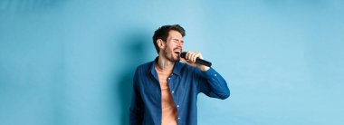 Kaygısız adam mikrofonla karaoke yapıyor, şarkı söylüyor, mavi arka planda duruyor. Boşluğu kopyala