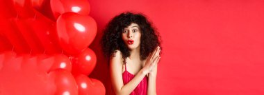 Heyecanlı kıvırcık saçlı güzel bir kadın, kalp balonlarının yanında duruyor ve avuç içlerini birbirine sürtüyor, iyi bir anlaşma ya da zevk bekliyor, kırmızı arka plan.