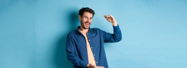 Neşeli modern adam gülümsüyor, büyük boy gösteriyor ve mutlu görünüyor, mavi arka planda duruyor. Boşluğu kopyala