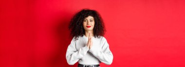 Namaste. Kıvırcık saçlı ve kırmızı dudaklı müteşekkir genç bir kadın minnettarlık gösteriyor. Kameraya gülümsüyor, stüdyo arka planında duruyor..