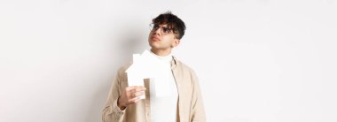 Emlak ve ipotek konsepti. Düşünceli genç adam yeni bir daire düşünüyor, elinde kağıt ev kesimi var ve sağ üst köşedeki logoya bak, beyaz arka planda duruyor..