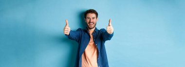 Yakışıklı, gülümseyen ve onay veren genç adam, mutlu bir yüzle evet de, mavi arka planda dur..