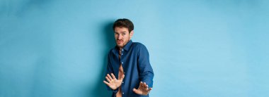 Çekingen genç adam uzak durmak istiyor, ellerini savunmaya uzatıp geri çekiliyor, iğrenç bir şeyi reddediyor, mavi arka planda duruyor..