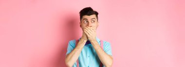Şok olmuş adam çenesini kapattı, ellerle dudaklarını kapadı ve soluna baktı. Pembe arka planda şaşkınlık içinde durdu..