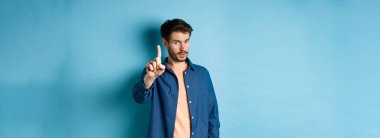 Genç bir adamın görüntüsü uyarı veriyor, ders veriyor, azarlamak için parmağını kaldırıyor, kameraya bakıyor, birine patronluk taslıyor, mavi arka planda duruyor..