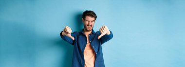 Tiksinç adam olumsuz görüş bildiriyor, baş parmaklarını aşağı ve dillerini gösteriyor, üzgün, kötü bir şeyi onaylamıyor, mavi arka planda duruyor..