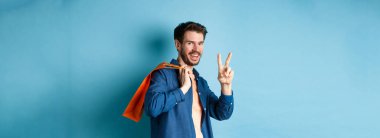 Omzunda turuncu alışveriş çantası tutan neşeli adam, gülümseyen ve barış işareti gösteren, mavi arka planda duran. Boşluğu kopyala