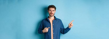 Tişörtlü yakışıklı genç adam, solu işaret ediyor, logoyu gösteriyor, mavi arka planda duruyor..