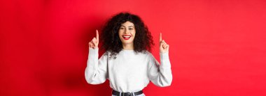 Kıvırcık saçlı mutlu genç bir kadın, parmaklarını havaya kaldırıp gülüyor, mükemmel beyaz bir gülümseme sergiliyor, kırmızı arka planda duruyor..