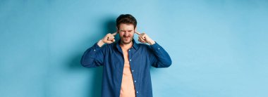 Sinirli genç adam sesi, parmaklarıyla kulaklarını kapatıyor ve yüzünü buruşturuyor, yüksek sesli müzikten rahatsız oluyor, mavi arka planda duruyor..