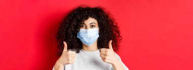 Covid-19 ve pandemik konsept. Yüz maskeli genç, kıvırcık bir kadının yakından çekilmiş fotoğrafı. Kırmızı arka planda duran güzel bir şey..