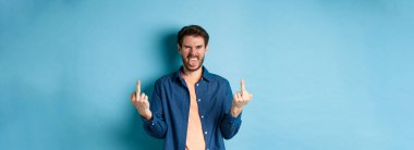 Ignorant and rude guy showing middle finger and smiling, say fuck you, standing on blue background. Copy space