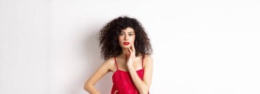 Güzellik ve moda. Şehvetli, kıvırcık saçlı, kırmızı dudaklı, zarif bir elbise giyen, yüzüne dokunan ve baştan çıkarıcı görünen beyaz bir kadın model..