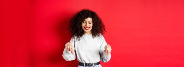 Kıvırcık saçlı ve kırmızı dudaklı modern bir kadın, parmaklarını eğip gülümsüyor ve boş bir alanda, stüdyo arka planında reklamlar yapıyor..