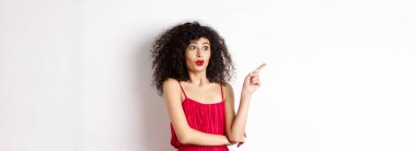 Kıvırcık saçlı heyecanlı kız, kırmızı elbise giyiyor, parmağını sola gösteriyor ve şaşırmış görünüyor, havalı reklamlar yapıyor, beyaz arka planda duruyor..