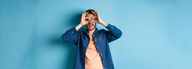 Komik beyaz adam el dürbününden bakıp gülümsüyor, ilginç bir şeye bakıyor, mavi arka planda şaşkın şaşkın duruyor..