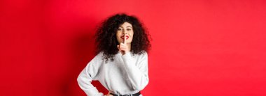 Kıvırcık saçlı ve makyajlı güzel, modern bir kadın, şşşş ve gülümsüyor, bir sır söylüyor, sürpriz yapıyor ve gülümsüyor, kırmızı arka planda duruyor..