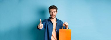Havalı adam baş parmağını gösteriyor ve elinde turuncu bir alışveriş çantası, mavi arka planda duruyor..