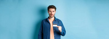Sakallı, sıradan kıyafetler giyen, plastik kredi kartı gösteren ve mavi arka planda kameraya bakan yakışıklı bir genç..