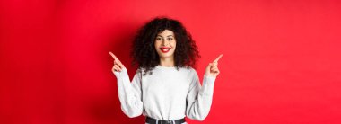 Çekici beyaz kadın yol gösteriyor, parmaklarını iki tanıtıma doğrultuyor, değişkenleri gösteriyor ve gülümsüyor, kırmızı arka planda duruyor..