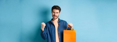 Yakışıklı gülümseyen adam evet diyor, alışveriş torbasını gösteriyor, iyi bir tanıtım teklifini kutluyor, mavi arka planda duruyor..