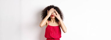 Heyecanlı kıvırcık saçlı, elbiseli, elleri ve parmaklarının arasından bakan, kameraya şaşırmış, beyaz arka planda duran.