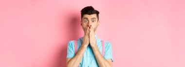 Şok olmuş bir adam bakıyor, elleriyle ağzını kapatıyor ve pembe arka planda durup kameraya bakıyor..