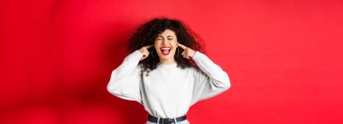 Sinirli kıvırcık kız parmaklarıyla kulaklarını tıkıyor ve bağırıyor. Kırmızı arka planda duran korkunç gürültüden rahatsız oluyor..