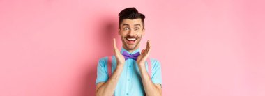 Mutlu genç adam şaşkınlık ve inançsızlıkla bakıyor, kameraya gülümsüyor, yüzün yanında el ele tutuşuyor, pembe arka planda papyon ve gömlekle duruyor..