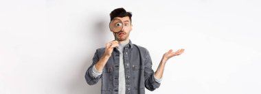 Kafası karışmış bir adam büyüteçle bakar, omuz silker, beyaz arka planda durur..