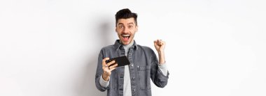 Heyecanlı adam cep telefonundan para kazanıyor ve seviniyor, elini kaldırıyor ve mutluluk ve neşe çığlıkları atıyor, beyaz arka planda akıllı telefonuyla duruyor..