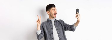 Akıllı telefondan selfie çeken neşeli bir gencin profili. Mobil uygulamayla fotoğraf çektirirken V işareti gösteriyor. Beyaz arka planda duruyor..