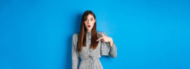 Şaşırmış genç bir kadın kendini inançsızlıkla işaret ediyor, seçiliyor ya da seçiliyor, mavi arka plana karşı elbise içinde duruyor..
