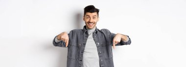 Heyecanlı, gülümseyen erkek manken parmaklarını aşağıya doğru gösteriyor, mutlu yüzlü logoyu gösteriyor, havalı bir promosyon sergiliyor, beyaz arka planda duruyor..