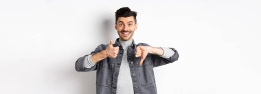 Mutlu gülümseyen adam ortalama bir hareket gösteriyor, baş parmağını kaldırıyor, karar veremiyor, bir şeyi orta derecede yargılıyor, beyaz arka planda duruyor..