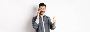Heyecanlı genç adam telefonda konuşuyor ve baş parmağını kaldırıyor, gülümsüyor, beyaz arka plana karşı duruyor..