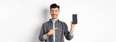 Yakışıklı gülümseyen adam akıllı telefon ekranını işaret ediyor, mutlu yüzlü cep telefonu görüntüsünü gösteriyor, beyaz arka planda duruyor..