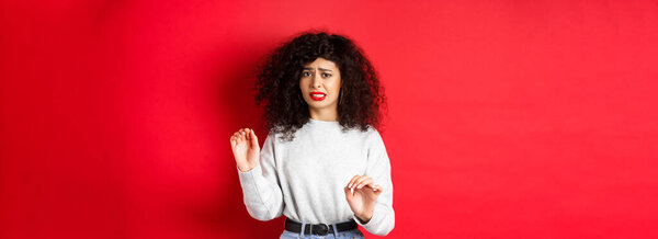 Отвратительная и неохотная женщина говорит нет, поднимая руки, чтобы заблокировать или отказаться, прося остановиться, выглядя неудобно, стоя на красном фоне.