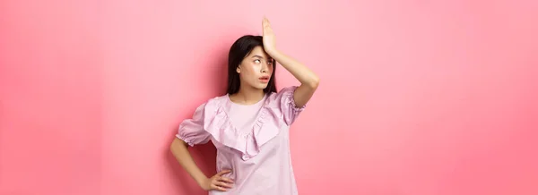 Annoyed asian girl roll eyes and facepalm, feeling tired and irritated with stupid conversation, standing against pink background.