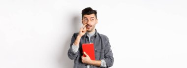 Adam burnunu karıştırıyor ve logoya bakıyor, elinde kırmızı kitap ya da planlamacı, beyaz arka planda duruyor..