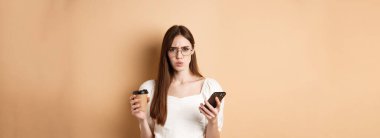 Kafası karışmış gözlüklü bir kız cep telefonu okuduktan sonra kameraya kaşlarını çatıyor, elinde kahve fincanı tutuyor, bej arka planda hoşnutsuz duruyor..